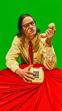 Young man stares at camera with serious expression, holding vintage phone receiver while seated at red table against green background. Concept of business, work-life balance. Pop art colors. Nostalgia clipart