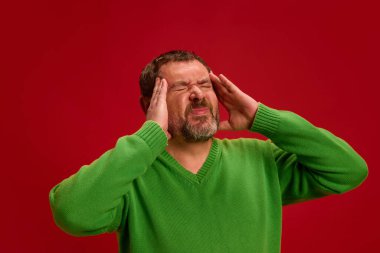 Olgun bir adam başını tutuyor ve acı çekiyor, migren, canlı kırmızı arka planda. Stres, tükenme. Sağlık, hastalık, virüs mevsimi, önleme kavramı. Ad