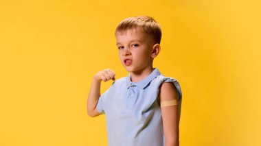 Küçük, kendine güvenen çocuk kaslarını gösteriyor ve sarı arka plandaki aşılar üzerine yamayla kolunu gösteriyor. Sağlık, koruma, dokunulmazlık, önleme kavramı.