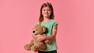 Elinde yamayla elinde birayla poz veren sevimli, mutlu küçük kız aşıdan sonra pastel pembe arka plana gülümsüyor. Sağlık, koruma, dokunulmazlık, önleme kavramı. Boşluğu kopyala.