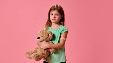 Elinde yamayla elinde birayla poz verip pastel pembe arka planda aşıdan korkan tatlı, korkmuş küçük bir kız. Sağlık, koruma, dokunulmazlık, önleme kavramı.