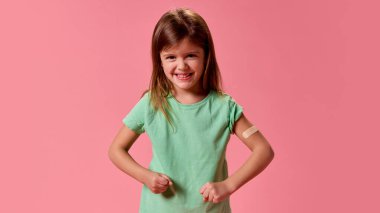 Pastel pembe arka planda aşıdan sonra elinde ilaç bandıyla poz veren virüslerle savaşmaya hazır küçük kız. Sağlık, koruma, dokunulmazlık, önleme kavramı.