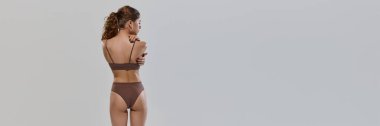 Banner, gri stüdyo arka planına karşı iç çamaşırlarıyla poz veren genç ve zayıf bir kadının arka görüntüsü. Doğal güzellik, kadın sağlığı, moda ve tarz kavramı. Ad