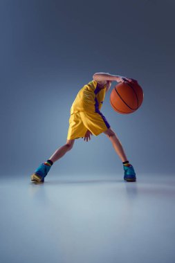 Genç bir çocuğun dikiz görüşü, canlı basketbol kıyafetleri, gri stüdyo arka planına karşı çalışma tekniği. Spor anlayışı, eğlence aktivitesi, hobi, rekabet. Ad