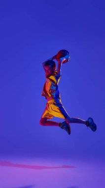 Oyuna odaklanmış, kendine güvenen genç basketbolcu gergin bir maçta neon ışıkta hareket ederek gradyan stüdyo arkaplanına doğru sıçrıyor. Spor anlayışı, eğlence aktivitesi, hobi, rekabet. Ad
