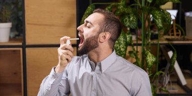 Genç adam, hasta, boğaz ağrısı semptomlarıyla ofisteki bu ağrıyı gidermek için sprey kullanıyor. Hastalık kavramı, sağlık sigortası, ilaç, ilaç. Ad.