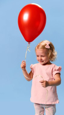 Küçük sevimli çocuk, pembe elbiseli kız, kırmızı balonu mavi stüdyo arka planında tutarken hafifçe gülümsüyor. Sağlık, koruma, dokunulmazlık, önleme kavramı.