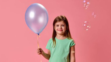 Mavi balon tutan, yeşil gömlek giyen ve elinde yamayla gülümseyen, pembe stüdyo arka planına karşı rahat poz veren bir kız. Sağlık, koruma, dokunulmazlık, önleme kavramı.