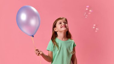 Neşeli bir ifadeyle yukarı bakan kız, elinde balonla, pembe stüdyo arka planında yüzen balonlarla çevrili. Sağlık, koruma, dokunulmazlık, önleme kavramı.