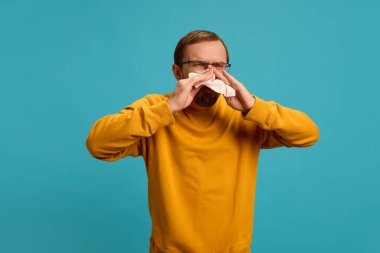 Hapşırmaktan acı çeken sıradan gözlük takan genç bir adam mavi stüdyo arka planına karşı burnunu peçeteye sümkürdü. Sağlık, ilaç, enfeksiyon ve tedavi kavramı. Ad.