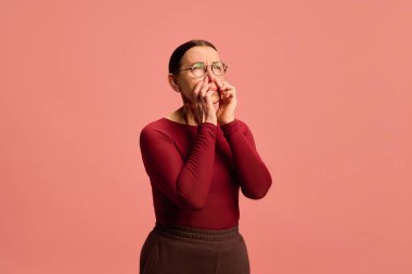 Burgundy bluzlu kadın iki eliyle burun köprüsünü tutuyor, şaşı gözler, pembe arka planda acı ya da rahatsızlık gösteriyor. Sağlık, ilaç, enfeksiyon ve tedavi kavramı. Ad.