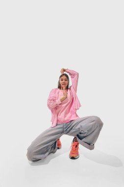 Kadın, pembe ve bol kot pantolon içinde dans ederken, beyaz stüdyo arka planına karşı eğlenceli bir şekilde diz çöker. Başarı kavramı, profesyonel gelişme, hareket. Ad