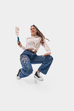 Beyaz gömlekli ve renkli kotlu genç bir kadın elinde boya silindiriyle öne eğilip beyaz stüdyo arka planına karşı yaratıcılığını ifade ediyor. Başarı kavramı, profesyonel gelişme, hareket. Ad