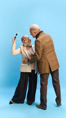 Kahverengi kareli ceketli adam ona doğru eğilirken, güneş gözlüğü kaldıran bir kadın mavi stüdyo arka planına doğru eğiliyor. Olgun insanlar ve moda kavramı. Ad
