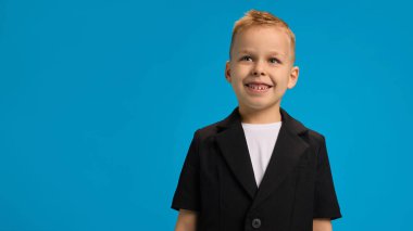Siyah ceket ve beyaz gömlek giyen gülümseyen çocuk, neşeli bir ifadeyle yukarı bakıyor, kendine güvenerek parlak mavi arka plana karşı duruyor. Çocukluk kavramı, güzellik, moda, tarz. Ad