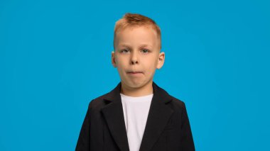 Siyah ceketli, beyaz gömlekli, dudakları hafifçe büzüşmüş parlak mavi arkaplanlı ciddi görünümlü bir çocuk. Çocukluk kavramı, güzellik, moda, tarz. Ad