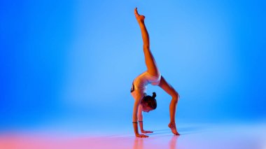 Genç, esnek, şenlikli kostümlü sanatçı kız mavi arka planda pembe neon ışıkta zor numaralar yapıyor. Profesyonel spor anlayışı, aktif yaşam tarzı, hobi. Ad