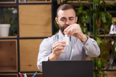 Genç adam iş yerinde oturur ve bir şişe burun spreyi açar. Mesajını eklemek için içinde negatif boşluk vardır. Seçici odaklı fotoğraf. Hastalık kavramı, sağlık sigortası, ilaç, ilaç. Ad.
