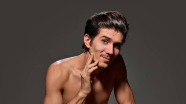 Elleri yanağına yakın bir adam neşeli bir gülümseme sergiliyor. Pürüzsüz saç stili ve koyu arkaplanda çıplak omuzları vardı. Erkek güzelliği ve yüz bakımı kavramı, yaşlanma karşıtı prosedürler.