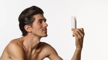 Kendine güvenen, bakımlı, beyaz şişeyi inceleyen, fotokopi alanı olan, odaklanmış bakışlı, beyaz arka planda göz hizasında tutan genç bir adam. Erkek güzellik anlayışı ve yüz bakımı, kozmetik.