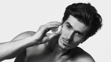 Siyah beyaz insan portresi, çıplak tişörtsüz, kameraya bakıyor, buz küpünü beyaz arka planda tutuyor. Erkek güzelliği ve yüz bakımı, cilt bakımı, kozmetoloji, cilt bakımı kavramı.