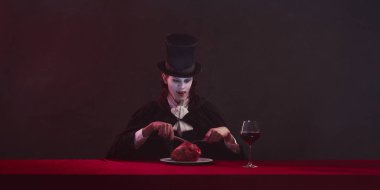 Korkunç bir adam, pelerinli ve silindir şapkalı vampir kırmızı masada oturuyor. Tabağında bir kadeh şarap ve kalp var. Ciddi bir ifadeyle ileri bakıyor. Tarihsel ve modernlik kavramı, yaratıcılık.