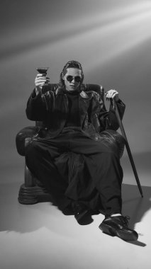 Deri ceketli ve güneş gözlüklü bir adamın siyah beyaz fotoğrafı. Sandalyede oturmuş kokteyl içerken vampire benziyor. Parti havası. Tarihi ve modernlik kavramı, Cadılar Bayramı Partisi.