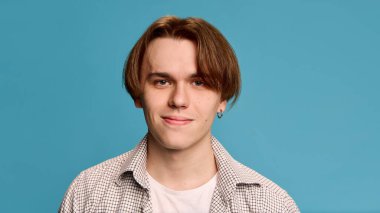 Genç adam ince gülümsüyor, ekose gömlek ve beyaz tişört giyiyor, açık mavi stüdyo arka planına karşı rahat bir poz veriyor. İnsan duyguları kavramı, kendini ifade etme. Ad