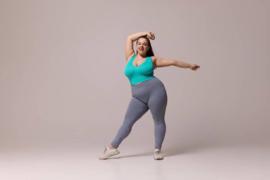 Mavi bluzlu ve taytlı tombul bir kadın gülümserken bir kolunu havaya kaldırıyor, dinamik bir poz veriyor, beyaz stüdyo arka planına karşı. Güzellik, zindelik, hareket, dans ve müzik kavramı.