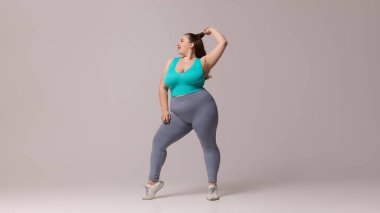 Mavi bluzlu, eli başının arkasında taytlı, beyaz stüdyo arka planında rahat duruşu olan, kendine güvenen bir kadın. Güzellik, zindelik, hareket, dans ve müzik kavramı.