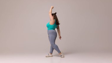 Mavi bluzlu şişman kadın ve geriye doğru uzanan taytlı beyaz stüdyo arka planına karşı enerjiyle zarif hareketler sergiliyor. Güzellik, zindelik, hareket, dans ve müzik kavramı.