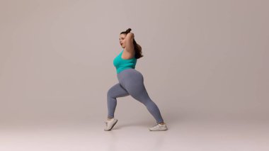 Mavi bluzlu ve taytlı bir kadın beyaz stüdyo arka planına karşı hamlesini yapıyor, gücünü ve dengesini kanıtlıyor. Güzellik, zindelik, hareket, dans ve müzik kavramı.