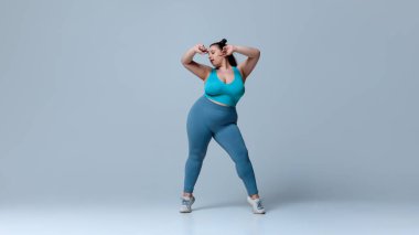 Artı mavi bluzlu ve taytlı kadın kollarını kaldırarak hareketli, beyaz stüdyo arka planına karşı neşeli hareketler sergiliyor. Güzellik, zindelik, hareket, dans ve müzik kavramı.