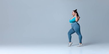 Mavi bluzlu ve taytlı şişman bir kadının yandan görünüşü beyaz stüdyo arka planına karşı enerji dolu bir şekilde öne çıkıyor. Güzellik, zindelik, hareket, dans ve müzik kavramı.