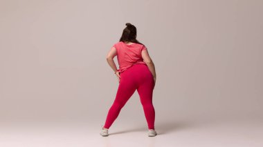 Kırmızı bluzlu ve taytlı genç şişman bir kadının arka plan fotoğrafı, kendine güvenen, minimalist arka planda kalçaların üzerinde el ele tutuşan. Güzellik, zindelik, hareket, dans ve müzik kavramı.