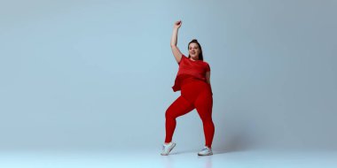 Kırmızı tişört giyen, kırmızı tayt giyen, beyaz spor ayakkabılı, elleri bej renkli stüdyo arka planında dans ederken gülümseyen tombul bir kadın. Güzellik, zindelik, hareket, dans ve müzik kavramı.