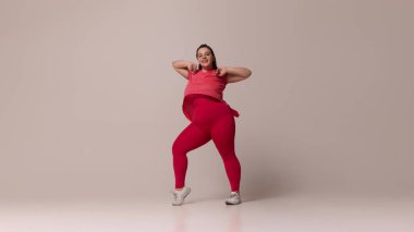 Dinamik bir portre. Mutlu gülümseyen, tombul bir kadın, kırmızı giyimli, beyaz stüdyo arka planında hip-hop dansı yapan. Güzellik, zindelik, hareket, dans ve müzik kavramı.