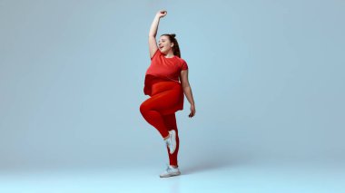 Kırmızı tişört giyen, kırmızı tayt giyen, beyaz spor ayakkabılı, elleri bej renkli stüdyo arka planında dans ederken gülümseyen tombul bir kadın. Güzellik, zindelik, hareket, dans ve müzik kavramı.