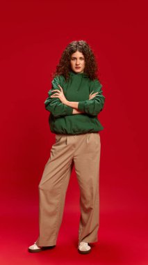 Uzun boylu, esmer, kıvırcık saçlı, el ele tutuşmuş, kırmızı arka planda kendine güvenen bir kadın portresi. İnsan duyguları, kendini ifade etme, moda ve tarz kavramı.