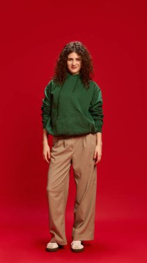 Genç, kıvırcık bir kadının, sıradan giyinmiş, kırmızı stüdyo arka planına hafifçe gülümseyen bir gülümsemeyle tam vücut fotoğrafı. İnsan duyguları, kendini ifade etme, moda ve tarz kavramı.