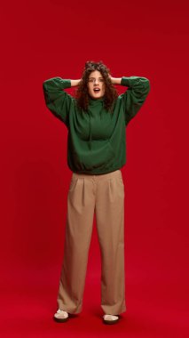 Kıvırcık saçlı, şık giyinmiş, kırmızı stüdyo arka planında hayret verici bir ifadeyle kameraya bakan genç bir kadın. İnsan duyguları, kendini ifade etme, moda ve tarz kavramı.