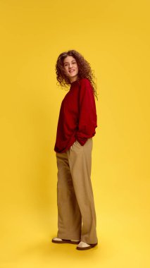 Çekici, gündelik kıyafetler giymiş, neşeli bir şekilde gülümseyen, güneşli sarı stüdyo geçmişine bakan bir kadın. İnsan duyguları, kendini ifade etme, moda ve tarz kavramı.