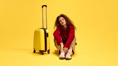 Güzel kıvırcık kadın bavulun yanında oturuyor, sarı stüdyo arka planında tatilleri bekliyor. İnsan duyguları, kendini ifade etme, moda ve tarz kavramı.