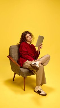 Kıvırcık saçlı, günlük kıyafetler giyen, tabletli sandalyede oturan ve sarı stüdyo geçmişine gülümseyen genç bayan. İnsan duyguları, kendini ifade etme, moda ve tarz kavramı.