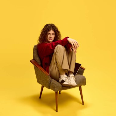 Kendine güvenen, çekici bir kadın, gündelik kıyafetler giymiş, sandalyeye oturmuş ve sarı arka planda kameraya bakıyor. İnsan duyguları, kendini ifade etme, moda ve tarz kavramı.