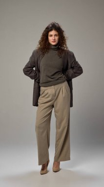Uzun boylu, esmer, kıvırcık saçlı, resmi giyinmiş, minimalist gri ayara poz veren çekici bir kadın fotoğrafı. İnsan duyguları kavramı, kendini ifade etme, moda, tarz.