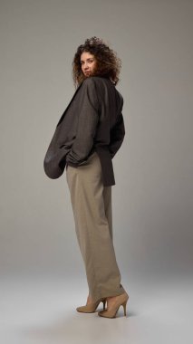 Şık giyimli, genç ve çekici bir kadın. Omuzlarının üzerinden bakıyor. Gri stüdyo geçmişine karşı güven gösteriyor. İnsan duyguları, kendini ifade etme, moda ve tarz kavramı.