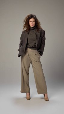 Gri stüdyo arkaplanına güvenle poz veren, resmi giyinmiş genç ve fit bir kadının vücut ölçülerinde fotoğrafı. İnsan duyguları, kendini ifade etme, moda ve tarz kavramı.