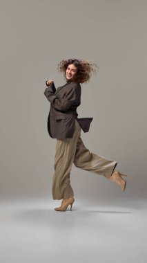 Genç ve çekici bir kadının dinamik çekimi, esmer kıvırcık saçlı, neşeli dans eden, gri stüdyo geçmişine karşı resmi kıyafetler giyen, insan duyguları kavramı, kendini ifade etme, moda ve tarz sahibi..