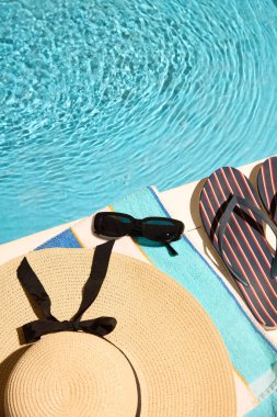 Siyah kurdeleli geniş kenarlı şapka, çizgili parmak arası terlikler, durgun havuz suyunun yanındaki renkli havluya yerleştirilmiş güneş gözlüğü yaz rahatlığının özünü yansıtıyor. Yaz tatili konsepti, turizm. Ad